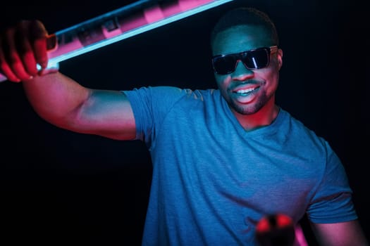 With sunglasses. Futuristic neon lighting. Young african american man in the studio.