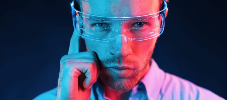 In glasses with display. Neon lighting. Young european man is in the dark studio.