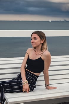 Young woman sitting on bench on wooden pier blurred beachside background. Attractive female enjoying the sea shore travel and active lifestyle concept. Springtime. Wellness wellbeing mental health inner peace Slow life digital detox