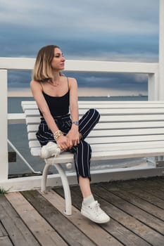 Young woman sitting on bench on wooden pier blurred beachside background. Attractive female enjoying the sea shore travel and active lifestyle concept. Springtime. Wellness wellbeing mental health inner peace Slow life digital detox