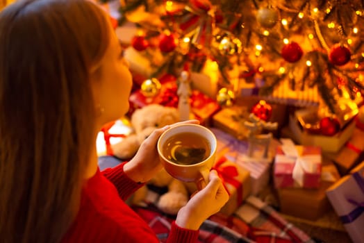 Young blond girl admiring beauty of christmas tree with a lot of gifts under it at home with cup of tea