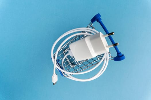 A phone charger in a supermarket cart on a blue background. Mini Shopping cart with Charger and Connecting Cable. top view