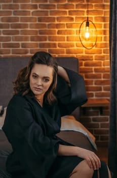 Portrait of a beautiful woman in a black bathrobe, who is sitting on the bed, looking at the camera.