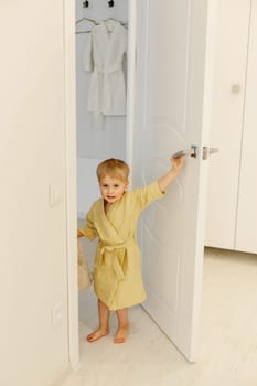 A boy in a yellow bathrobe stands at the entrance to the bathroom, smiling.