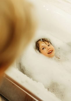Happy little girl swims in a bathtub full of foam. Looks at mom.