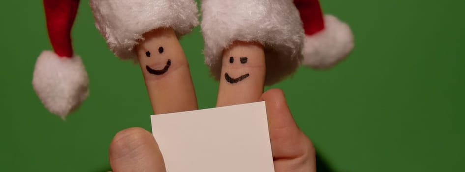 Fingers dressed in Santa-Claus red-white hats. Two happy finger smileys faces holding empty paper note for your text copy space against green background. Happy family celebrating concept for Christmas or New Years day. Finger couple Advertisement