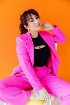 a woman in bright suit sits on a chair posing on an orange background