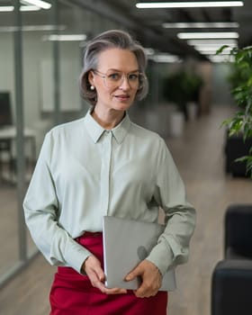 Mature caucasian woman stands with a laptop among the office