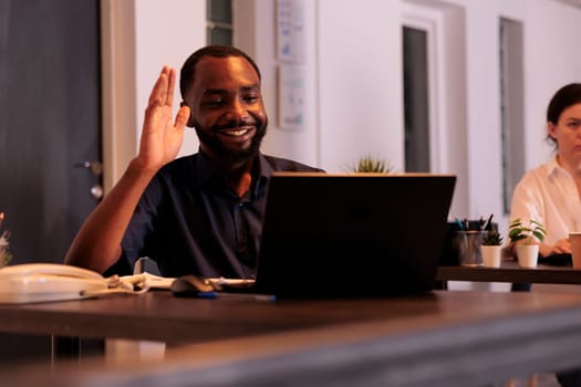 Smiling employee talking on videoconference with teamlead, waving hi, corporate worker answering teleconference call in office at night. African american man remote conversation in coworking space