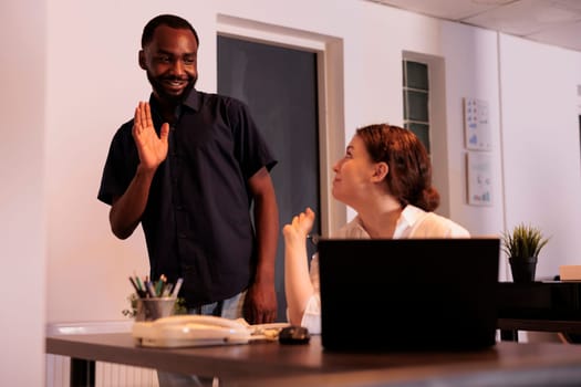 Office manager greeting new colleague, waving hi in coworking space, woman working on laptop, talking with coworker. Smiling african american man meeting employee, workers communication