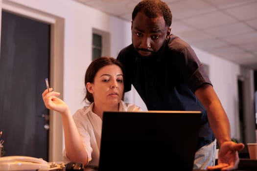 Coworkers reading company report on laptop together, project manager and employee working tohether in office at night. Diverse corporate workers checking report on computer in coworking space