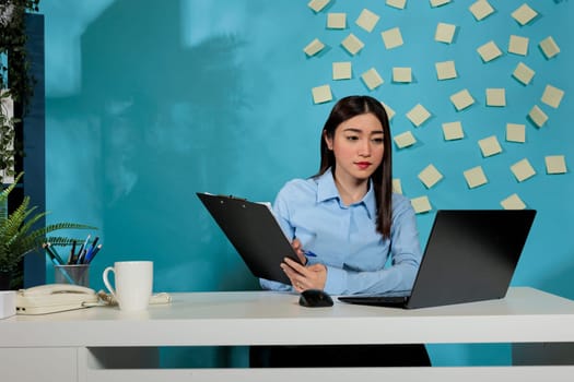 Female corporate assistant reviewing task list on laptop computer and taking notes on clipboard. Concentrated professional woman working from modern office sitting at desk.