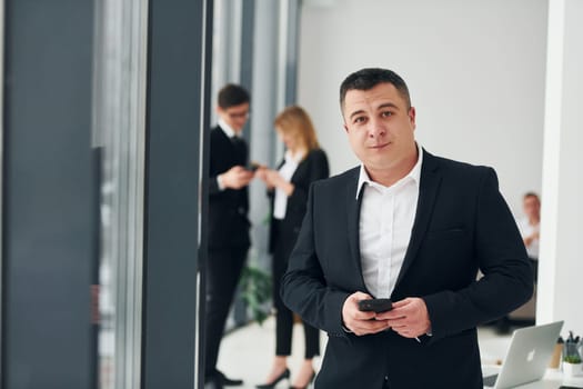 Man holds phone. Group of people in official formal clothes that is indoors in the office.