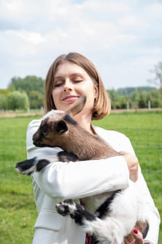 Young woman plays with goat kids, feeding them, sun shining over farm in background, High quality photo