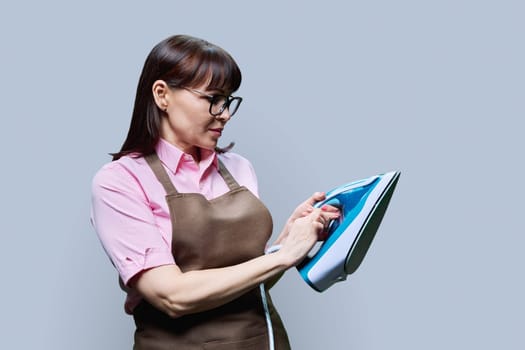 Woman in an apron posing with an iron, on a gray background. Female housewife, dry cleaning laundry worker, lifestyle work staff service concept