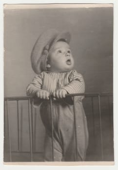 THE CZECHOSLOVAK SOCIALIST REPUBLIC - CIRCA 1960s: Vintage photo shows a small cute boy stands in the cot. He wears a flat cap. Retro black and white photography. Circa 1970s.