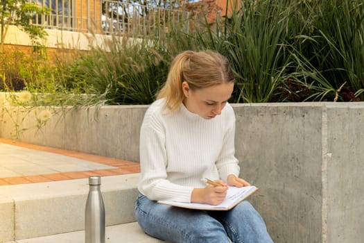 Young student study with notebook in park. Drinking water, hot tea or coffee from reusable metal bottle. Writing gratitude journal self reflection self discovery Outdoors warm autumn seashore.