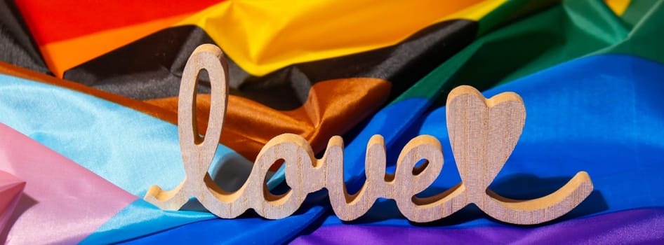 Banner Wooden word love with heart romance on Rainbow LGBTQIA flag made from silk material. Valentine's Day greeting card. Symbol of LGBTQ pride month. Equal rights. Peace and freedom. Support LGBTQIA community. Diversity equality