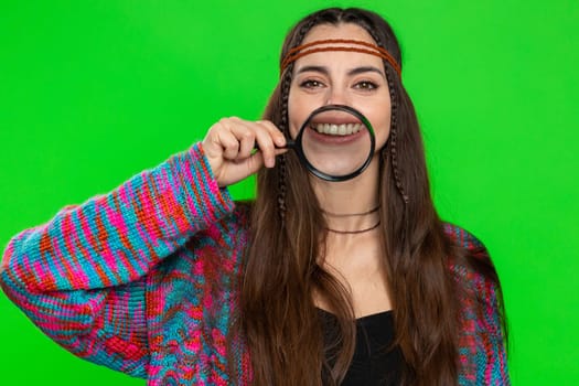 Young woman holding magnifier glass loupe on healthy white teeth, looking at camera with happy expression, showing funny silly face smiling mouth. Pretty hippie girl isolated on chroma key background