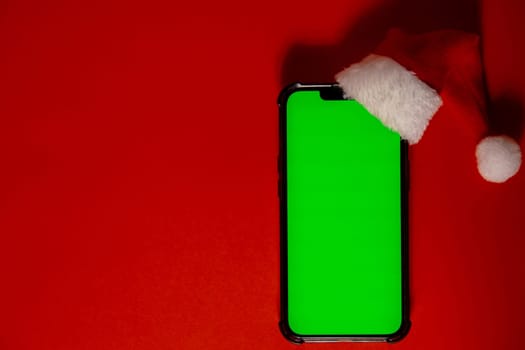 Mobile phone dressed in Santa-Claus red-white hat with chroma key screen against red background. Concept for Christmas or New Years holidays. Blank cell phone. Digital gadget, technology copy space wireless wishlist concept. Social media advertisement