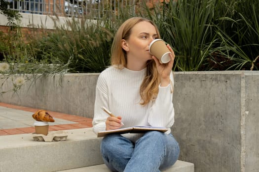 Young student study with notebook in park. Drinking coffee and eat croissants. Writing gratitude journal self reflection self discovery Outdoors warm autumn seashore.