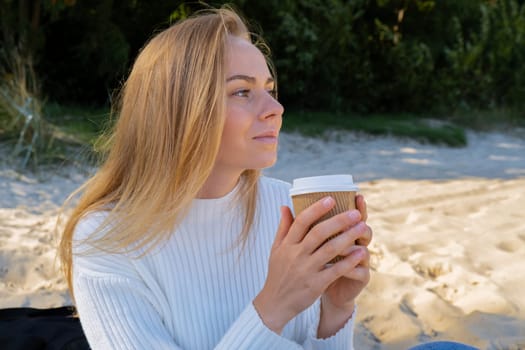 Happy young woman drinking coffee from paper cup takeaway on the beach sea ocean. Wearing wireless headphones doing audio healing sound therapy being mindful Leisure in nature. Wellbeing unity with nature health mindfulness. Enjoy outdoor lifestyle relaxation