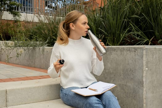 Young student study with notebook in park. Drinking water, hot tea or coffee from reusable metal bottle. Writing gratitude journal self reflection self discovery Outdoors warm autumn seashore.