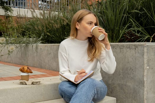 Young student study with notebook in park. Drinking coffee and eat croissants. Writing gratitude journal self reflection self discovery Outdoors warm autumn seashore.