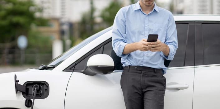 Progressive businessman talking on the phone, leaning on electric car recharging with public EV charging station, apartment condo residential building on the background as green city lifestyle.