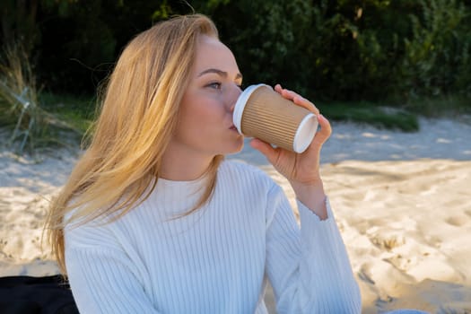 Happy young woman drinking coffee from paper cup takeaway on the beach sea ocean. Wearing wireless headphones doing audio healing sound therapy being mindful Leisure in nature. Wellbeing unity with nature health mindfulness. Enjoy outdoor lifestyle relaxation