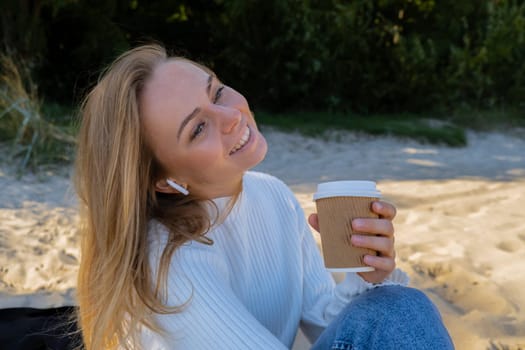 Happy young woman drinking coffee from paper cup takeaway on the beach sea ocean. Wearing wireless headphones doing audio healing sound therapy being mindful Leisure in nature. Wellbeing unity with nature health mindfulness. Enjoy outdoor lifestyle relaxation
