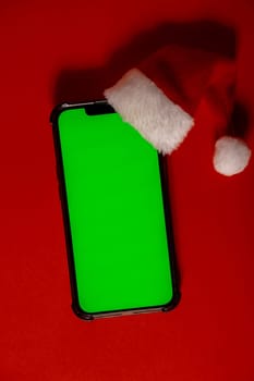 Mobile phone dressed in Santa-Claus red-white hat with chroma key screen against red background. Concept for Christmas or New Years holidays. Blank cell phone. Digital gadget, technology copy space wireless wishlist concept. Social media advertisement