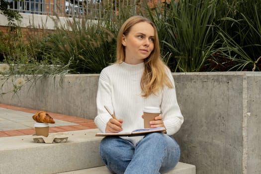 Young student study with notebook in park. Drinking coffee and eat croissants. Writing gratitude journal self reflection self discovery Outdoors warm autumn seashore.