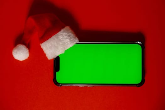 Mobile phone dressed in Santa-Claus red-white hat with chroma key screen against red background. Concept for Christmas or New Years holidays. Blank cell phone. Digital gadget, technology copy space wireless wishlist concept. Social media advertisement