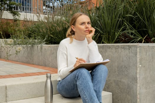 Young student study with notebook in park. Drinking water, hot tea or coffee from reusable metal bottle. Writing gratitude journal self reflection self discovery Outdoors warm autumn seashore.