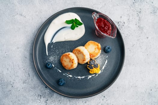 Cottage cheese pancakes, syrniki on plate with sour cream and jam, blueberry and edible flowers. Healthy and delicious morning breakfast.