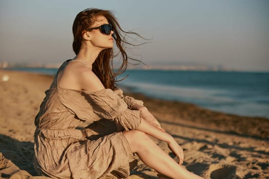 red-haired woman in dark sunglasses and sand-colored dress on the beach enjoying the sunset. High quality photo