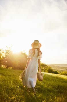a woman in a long light dress walks through the countryside in a hat and with a basket in her hands in the rays of the setting sun enjoying nature. Photo from the back. High quality photo