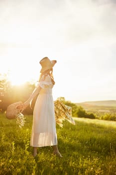 a woman in a long light dress walks through the countryside in a hat and with a basket in her hands in the rays of the setting sun enjoying nature. High quality photo