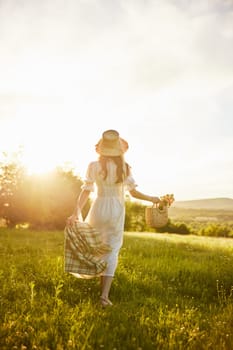 a woman in a long light dress walks through the countryside in a hat and with a basket in her hands in the rays of the setting sun enjoying nature. High quality photo