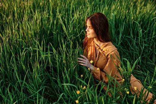 a happy woman is lying in the tall green grass in a long orange dress and smiling pleasantly looks away holding the leaves with her hand, enjoying nature. High quality photo