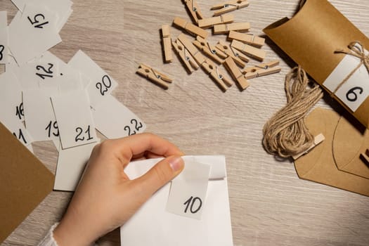 Unrecognizable young woman sticks number on craft bag, fastens with clothespin. Female making kraft paper for homemade advent calendar Made with your own hands step by step DIY crafts do it yourself. Preparation to christmas concept. Seasonal activities for children family winter holidays. Eco friendly presents gifts. open the package every day