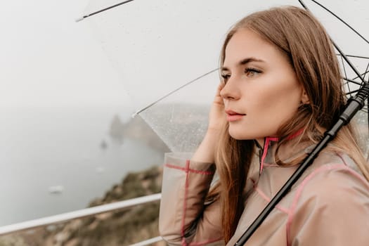 Woman rain park. Happy woman portrait wearing a raincoat with transparent umbrella outdoors on rainy day in park near sea. Girl on the nature on rainy overcast day