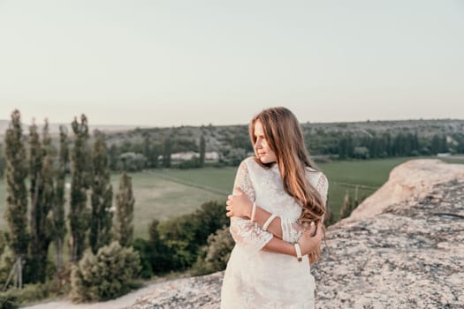 Romantic beautiful bride in white dress posing with sea and mountains in background. Stylish bride standing back on beautiful landscape of sea and mountains on sunset