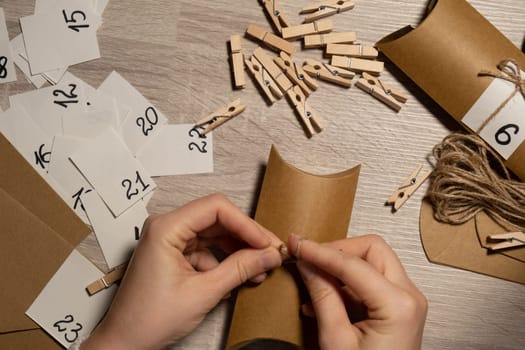 Unrecognizable young woman sticks number on craft bag, fastens with clothespin. Female making kraft paper for homemade advent calendar Made with your own hands step by step DIY crafts do it yourself. Preparation to christmas concept. Seasonal activities for children family winter holidays. Eco friendly presents gifts. open the package every day