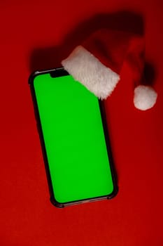 Mobile phone dressed in Santa-Claus red-white hat with chroma key screen against red background. Concept for Christmas or New Years holidays. Blank cell phone. Digital gadget, technology copy space wireless wishlist concept. Social media advertisement