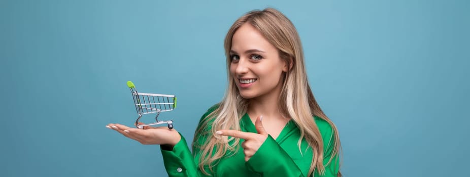 stylish european woman shopper in green shirt with supermarket trolley on blue studio background.