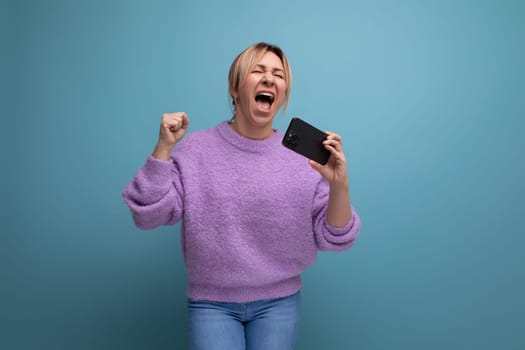 energetic blond young woman consultant in purple hoodie holding smartphone with mockup and singing on blue background with copy space.