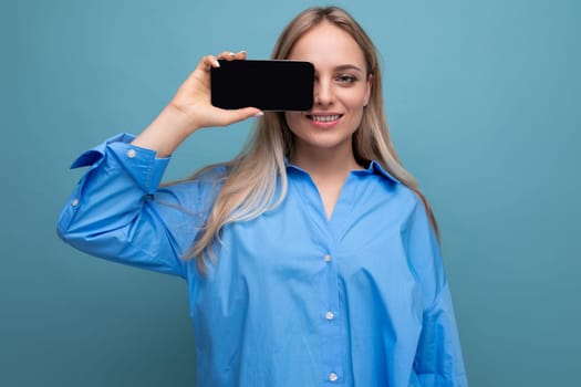 smiling informal blond girl holding smartphone with blank screen mockup for website on blue background.