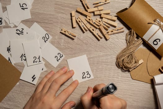 Unrecognizable young woman sticks number on craft bag, fastens with clothespin. Female making kraft paper for homemade advent calendar Made with your own hands step by step DIY crafts do it yourself. Preparation to christmas concept. Seasonal activities for children family winter holidays. Eco friendly presents gifts. open the package every day